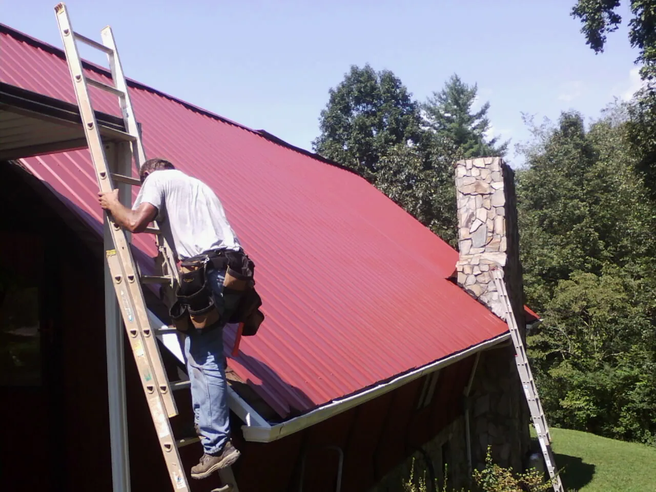 Roofer on a ladder beside a red metal roof and stone chimney — Soffit & Fascia Repair by Roofing Brothers in Luthersburg